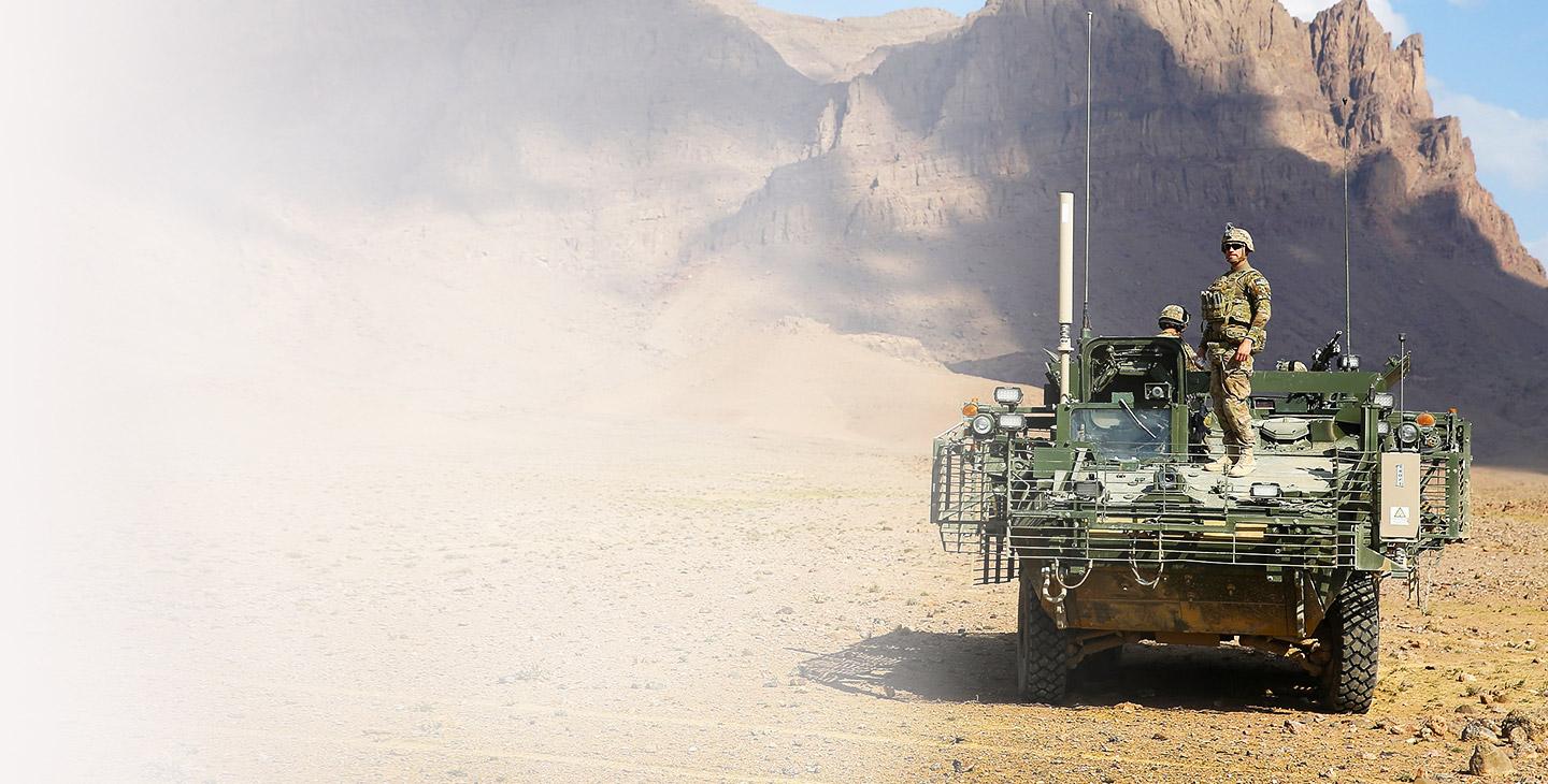 一名身穿战斗装备的军人站在一辆绿色军车的前部，背景是岩石山脉