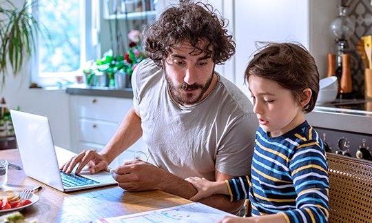 一个男人坐在餐桌前的笔记本电脑前, 在他儿子旁边，儿子正在给他看纸上的东西